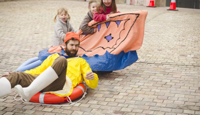 An outdoor Boats production photo. A Polyglot artist in a yellow raincoat, brown trousers, white gumboots, and orange hat sits in an orange flotation ring in a sandstone courtyard. Behind him are three children in a a pale orange and blue vessel, looking at the artist interestedly.