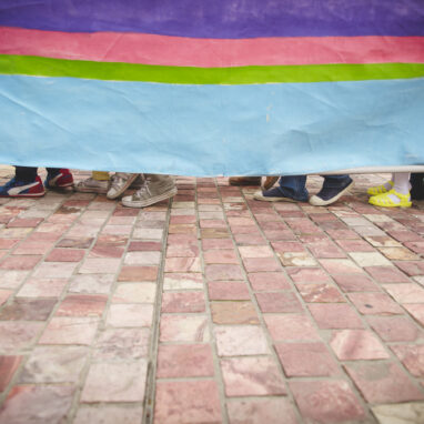 A Boats production photo. A rainbow vessel obscures eight people inside of it, who are walking in the same direction on a brick path. Only their shoes are visible.