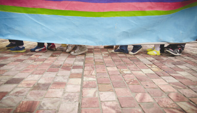 A Boats production photo. A rainbow vessel obscures eight people inside of it, who are walking in the same direction on a brick path. Only their shoes are visible.