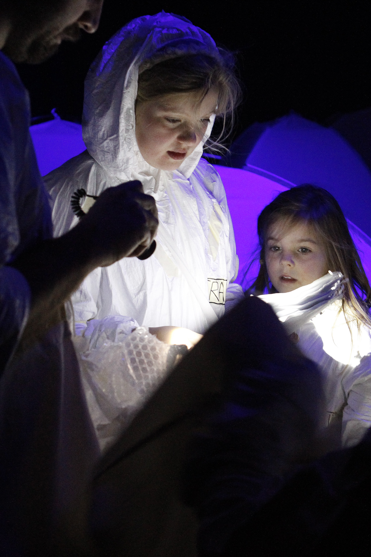 A Separation Street production photo. Two children wearing white boiler suits stand in a darkened space, looking at something illuminated by a torch/flashlight. Purple light illuminates the background.