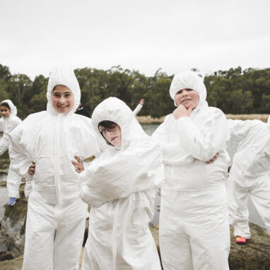 A school residency photo. Five students wearing boiler suits pose for the camera on rocks next to a body of water. There are trees and grey sky in the background.
