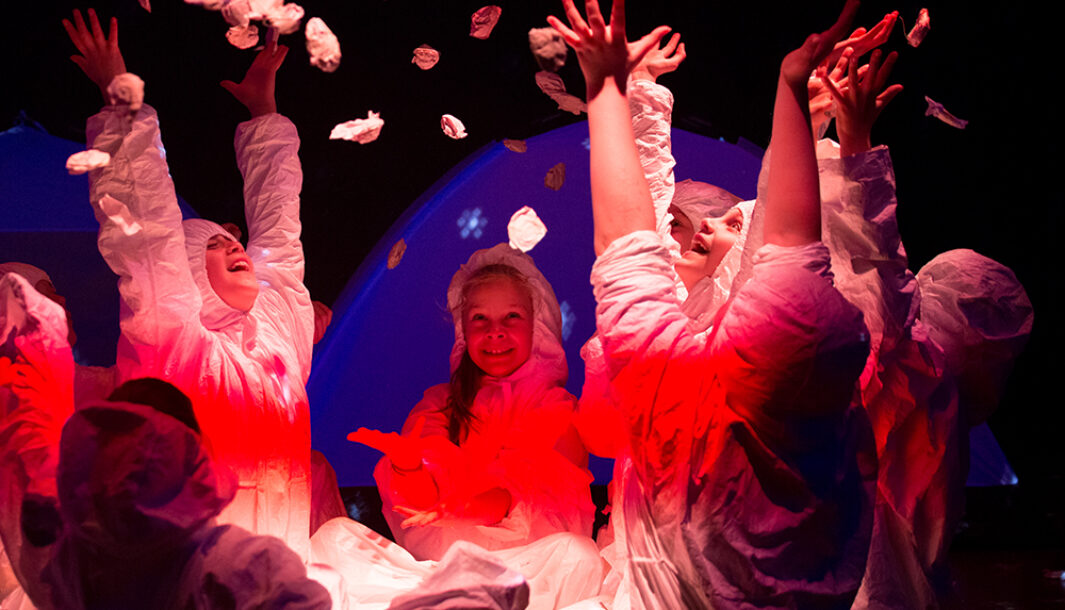 A Separation Street production photo. Children sit in a circle, their bodies illuminated by a red light. They throw paper petals into the air. Photo by Greta Costello.