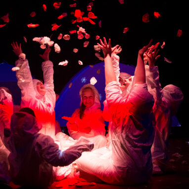 A Separation Street production photo. Children sit in a circle, their bodies illuminated by a red light. They throw paper petals into the air. Photo by Greta Costello.