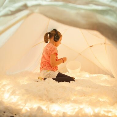 A Voice Lab production photo. A child sits on their legs in an softly lit dome. They are wearing headphones. Photo: Sarah Walker.