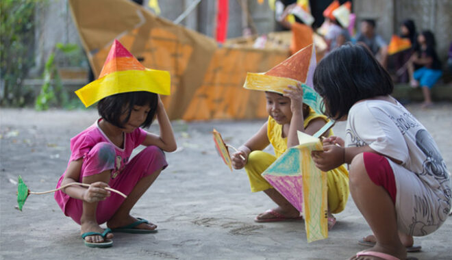A creative development photo from Indonesia. Three small children crouch on grey concrete, wearing or holding colourful, handmade boat-shaped hats. A large paper boat and other people are visible in the background.