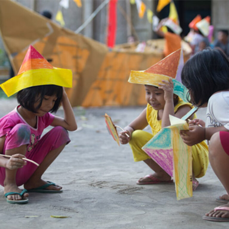 A creative development photo from Indonesia. Three small children crouch on grey concrete, wearing or holding colourful, handmade boat-shaped hats. A large paper boat and other people are visible in the background.