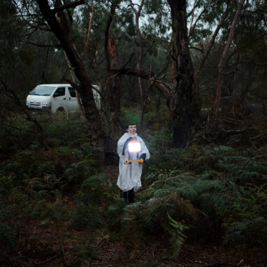 A school residency performance photo. A child in a white rain poncho, holding an illuminated lantern, stands in the Australian bush, surrounded by green shrubs and trees. They are speaking to an audience out of frame. A white van is parked in the distance behind them.