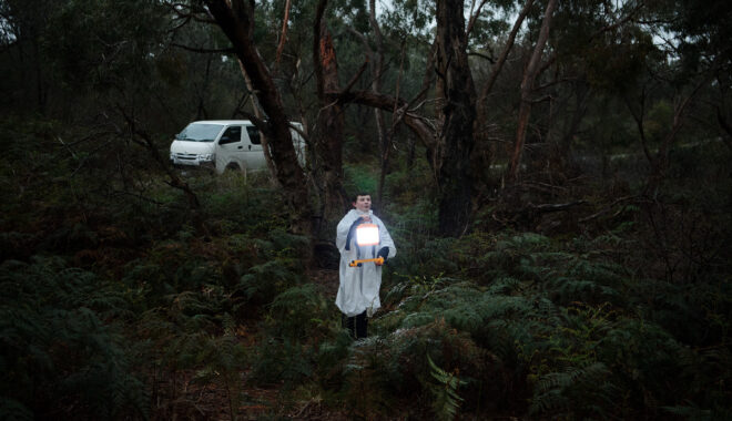 A school residency performance photo. A child in a white rain poncho, holding an illuminated lantern, stands in the Australian bush, surrounded by green shrubs and trees. They are speaking to an audience out of frame. A white van is parked in the distance behind them.