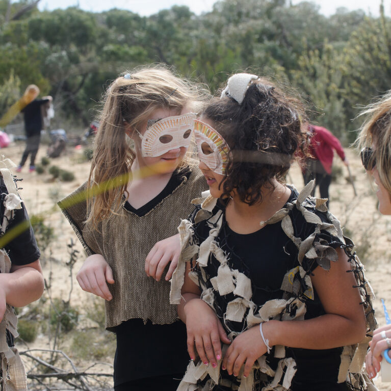 A Mahogany Rise residency photo. A group of children wearing handmade paper costumes standing in the bush with an artist. Other students and artists are visible in the background.