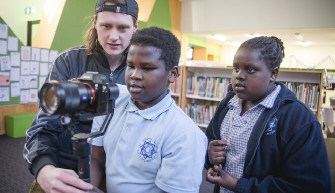 A 5678 Film Club photo. Two students and a Polyglot artist stand behind a camera in a school library. They are all looking at the camera intently.