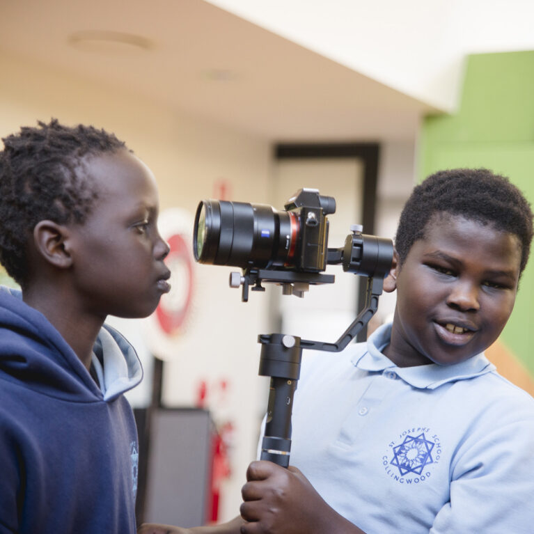 A 5678 Film Club photo. Two students stand in a school library. One holds a camera and is looking directly at the photographer. The other is side on, looking into the camera the other is holding.