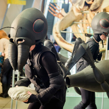 An Ants production photo. Polyglot artists in intricate black Ant costumes move through a large museum foyer, holding giant crumbs. An enormous skeleton and families visiting the museum are visible in the background. Photographer: Theresa Harrison