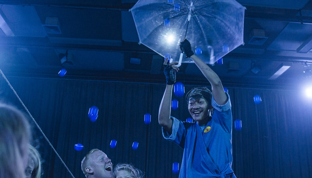 A Cerita Anak production photo. A performer shields themselves from falling blue balls with a blue umbrella. Photo: Theresa Harrison.