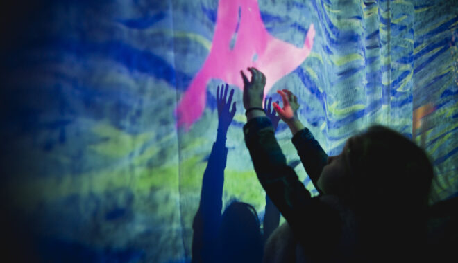A Cerita Anak production photo. A small child in silhouette reaches a hand towards an illustration of a pink sea creature amongst blue and green ocean water projected onto a screen.