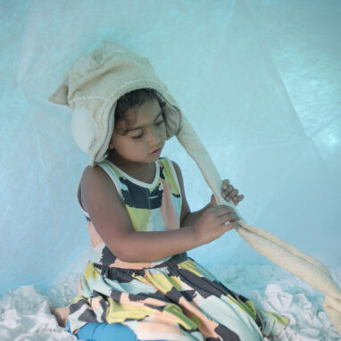 A Voice Lab production photo. A child sits in a soft, white, enclosed space that is illuminated with blue light. They wear a soft fabric 'crown' with a long cord, which they hold with both hands.