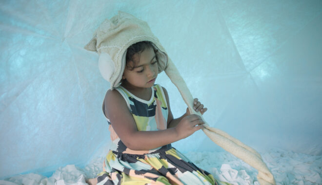 A Voice Lab production photo. A child sits in a soft, white, enclosed space that is illuminated with blue light. They wear a soft fabric 'crown' with a long cord, which they hold with both hands.