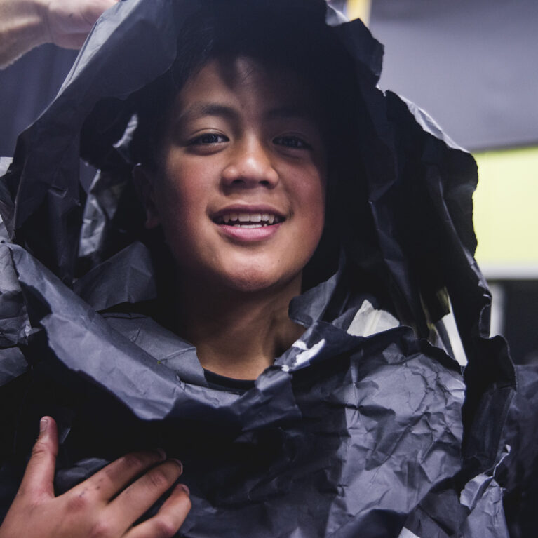 A Mahogany Rise school residency photo. A child wrapped in black paper smiles at the camera.
