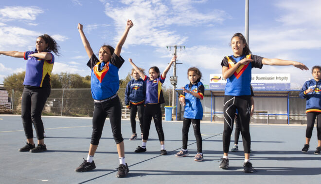 A First On The Ladder photo. A group of children in blue Rumbalara sports jerseys choreograph a dance on a netball court. Blue sky with sparse white clouds is visible behind them.