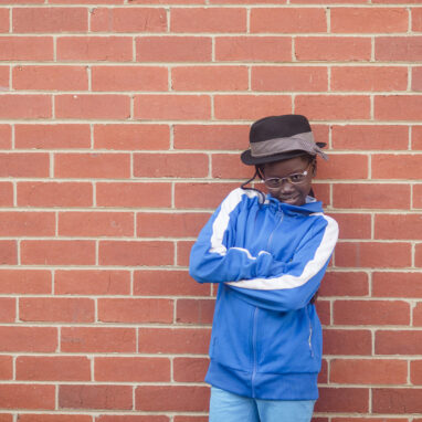 A 5678 Film Club photo. A student in a black hat, blue jumper and glasses stands in front of a red-brick wall. Their arms are crossed and they are smiling at the camera.