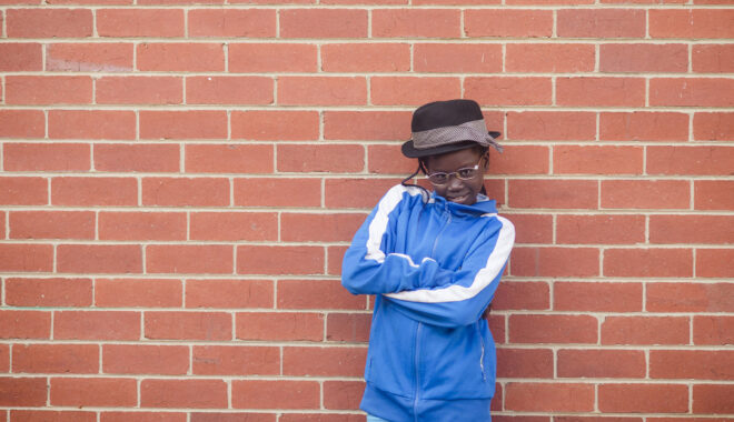 A 5678 Film Club photo. A student in a black hat, blue jumper and glasses stands in front of a red-brick wall. Their arms are crossed and they are smiling at the camera.
