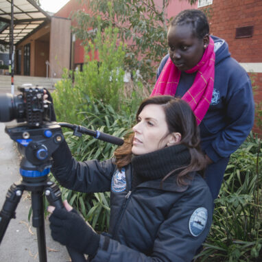 A 5678 Film Club photo. A Polyglot artist and a student stand behind a camera on a tripod, watching footage back.