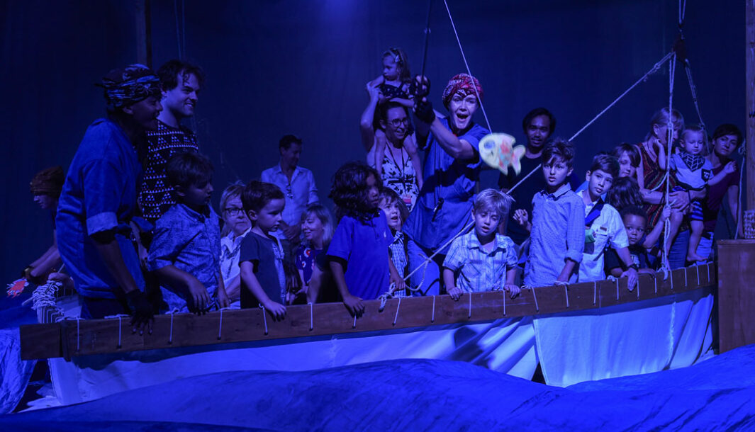 A Cerita Anak production photo. A makeshift boat holds children and performers. They are looking into an ocean constructed from billowing silk cloths. A performer flings a fishing rod with a paper attached to it into the ocean. They are illuminated by soft down-cast lighting.