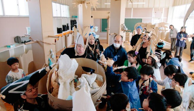 A Paper Planet workshop photo. Children and Polyglot artists wearing various paper costumes crowd around a large cardboard structure. They are in a classroom filled with tall brown cardboard trees.
