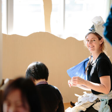 A Paper Planet production photo. A Polyglot artist wearing a handmade paper costume kneels on the ground amidst tall brown cardboard trees. She has turned her head and is smiling at the camera. Other people are visible around her.