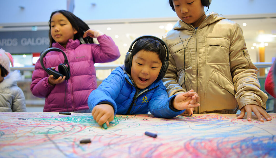 A Sound of Drawing production photo. Two children wearing headphones stand at a table covered in brown paper, drawing with pastels. A third child stands near them, holding a pair of headphones. Photographer: Sarah Walker.