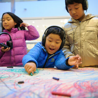 A Sound of Drawing production photo. Two children wearing headphones stand at a table covered in brown paper, drawing with pastels. A third child stands near them, holding a pair of headphones. Photographer: Sarah Walker.