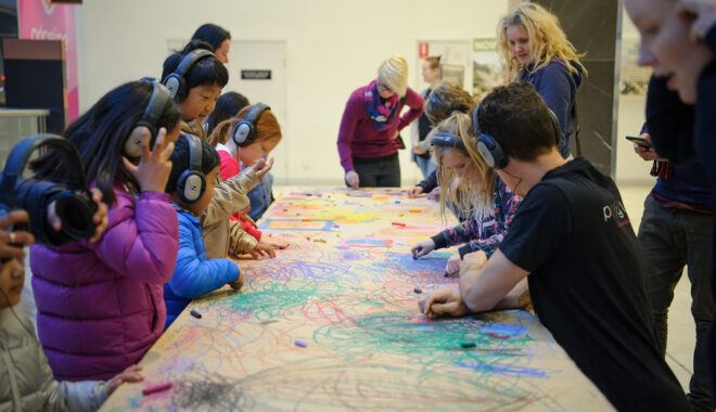 A Sound of Drawing production photo. A long table, covered in brown paper and filled with colourful drawing, is surrounded by children and adults. Some are wearing headphones and most are drawing.