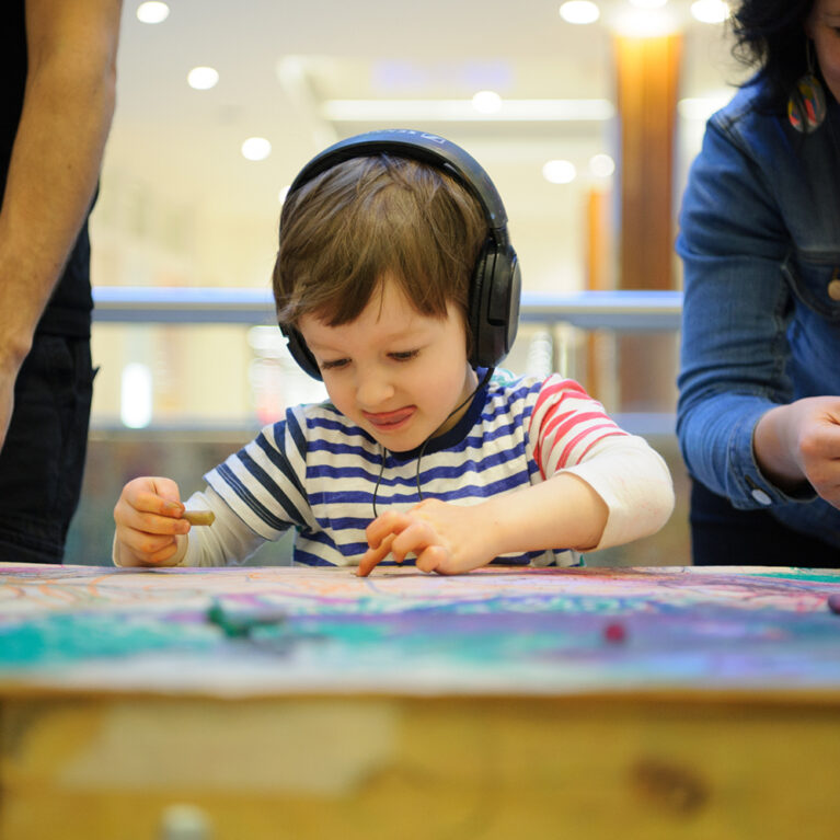 A Sound of Drawing production photo. A child with dark hair, wearing black headphones and a striped t-shirt, draws intently on a table covered in brown paper. Adults stand on either side of him.
