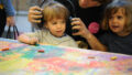 A Sound of Drawing production photo. Two children stand at a table covered in paper filled with colourful drawing. A Polyglot artist places a pair of black headphones on the child closest to the table.