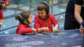 A Sound of Drawing production photo. Two children wearing red jumpers and black headphones stand together at a table covered in black paper. They are smiling and talking together, while drawing with chalk.