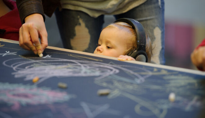 A Sound of Drawing production photo. A very small child wearing headphones stands at a table covered in black paper - they can only just see over it. Their adult standing behind them draws on the paper with chalk.