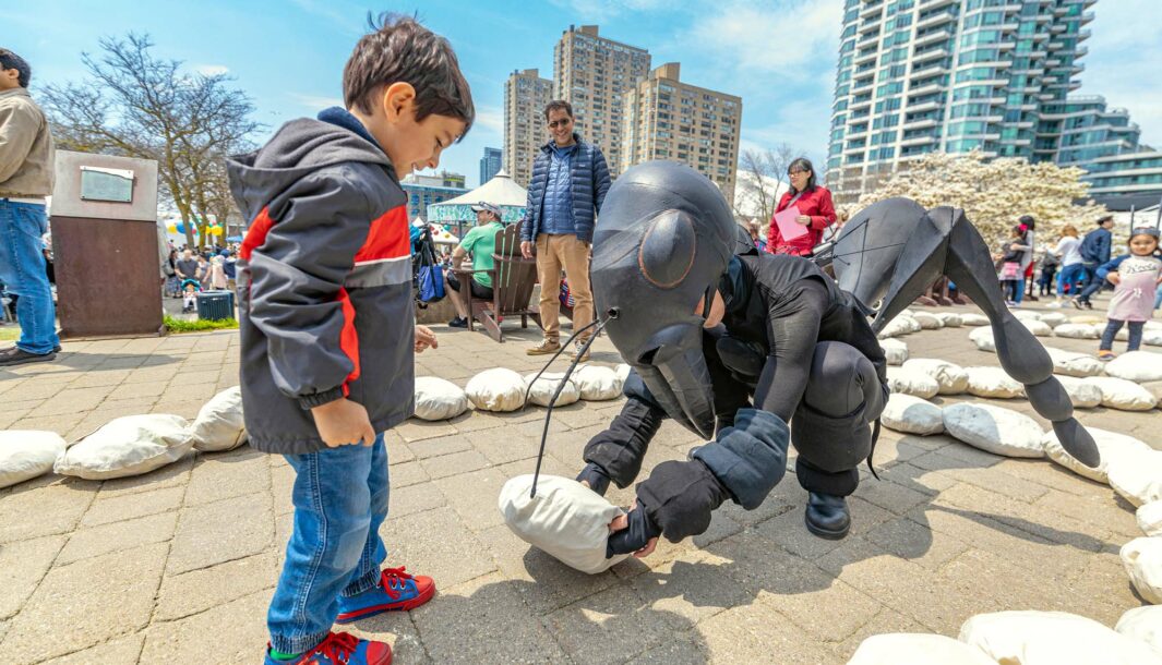 An outdoors Ants production photo. A Polyglot artist in an intricate Ant costume crouches in front of a child, offering them a giant breadcrumb. They are surrounded by lines of crumbs, and blue sky and tall buildings are in the background.