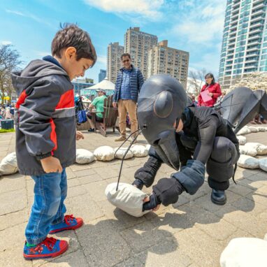 An outdoors Ants production photo. A Polyglot artist in an intricate Ant costume crouches in front of a child, offering them a giant breadcrumb. They are surrounded by lines of crumbs, and blue sky and tall buildings are in the background.