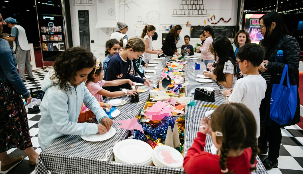 A Feast production photo. Children and families stand around long tables covered in black-and-white checked tablecloths, creating and playing with colourful paper, cardboard, cellophane and glue. Photographer: Jason Lau, courtesy of Arts Centre Melbourne