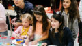 A Feast production photo. Four children and an adult are clustered at a table, making creations on paper plates.