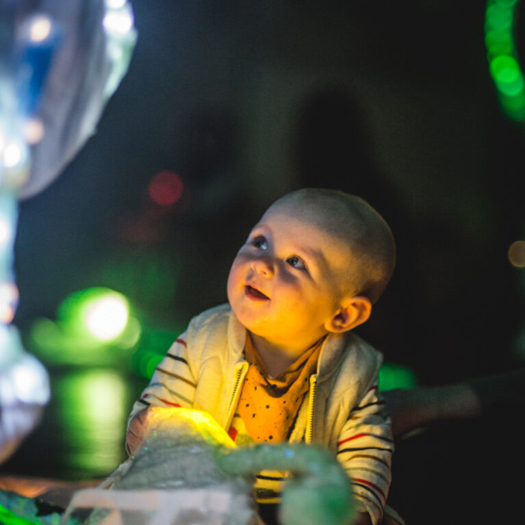 A Light Pickers production photo. A baby sits on the floor in a darkened space, surrounded by abstract glowing objects. They gaze upwards, fascinated.