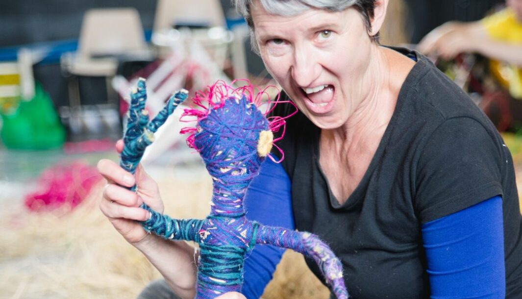 A Manguri Wiltja production photo. A person with short hair smiles and holds a doll made from purple woven string. The doll has woven purple arms, a purple head, a blue body and pink hair.
