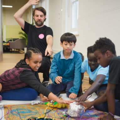 A Clippy workshop photo. A group of children sit on the floor, reaching towards a colourful object connected to large balls of aluminium foil. An excited Polyglot artist in a black t-shirt kneels behind them, one arm raised, the other pointing at the object.