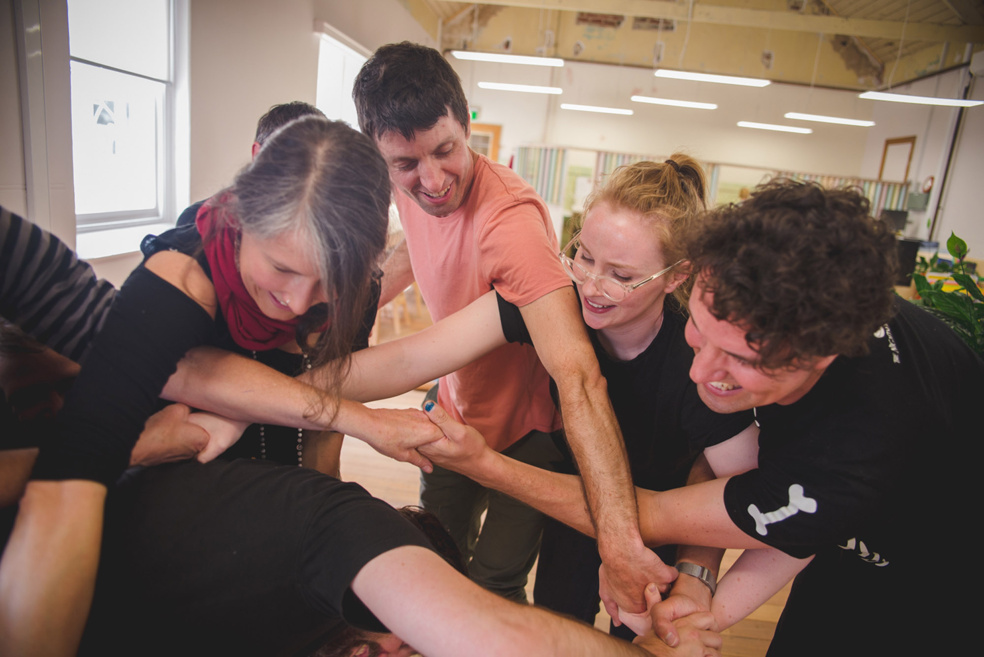 A professional development workshop photo. A group of artists have tangled their arms together, and are trying to untangle themselves without letting go of each other's hands.