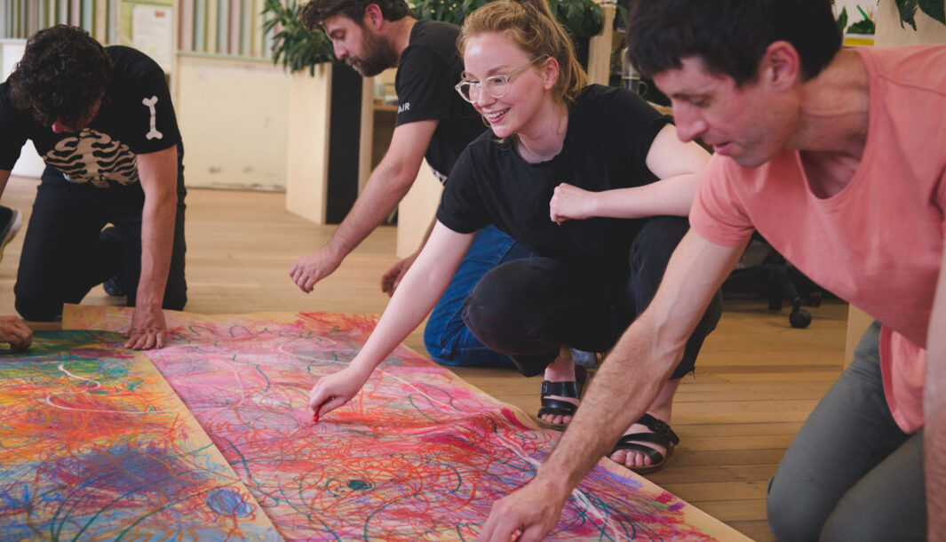 A professional development workshop photo. A group of artists crouch around a large piece of paper on the floor, drawing with coloured pastels.