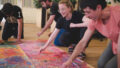 A professional development workshop photo. A group of artists crouch around a large piece of paper on the floor, drawing with coloured pastels.