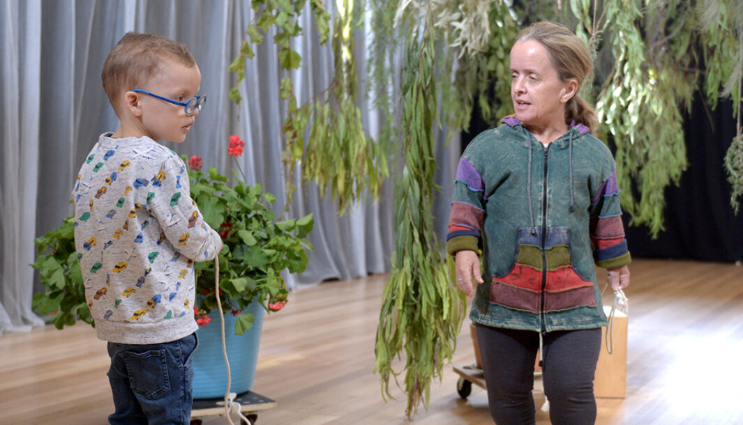 A Parked development photo. An adult and a child interact with one another in a well-lit theatre space with wooden floors under a hanging gum tree branch. Photo: Carla Gottjens.