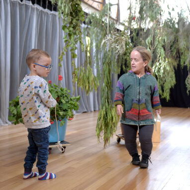 A Parked development photo. An adult and a child interact with one another in a well-lit theatre space with wooden floors under a hanging gum tree branch. Photo: Carla Gottjens.