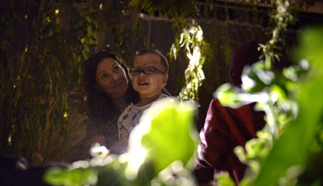 A Parked creative development photo. An adult and child stand together amidst green foliage, illuminated by dramatic theatre lighting.