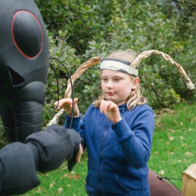 An outdoor Ants production photo. A Polyglot artist in an intricate Ant costume interacts with a child wearing a blue jumper and handmade paper antennae. Green grass and shrubs are visible in the background.