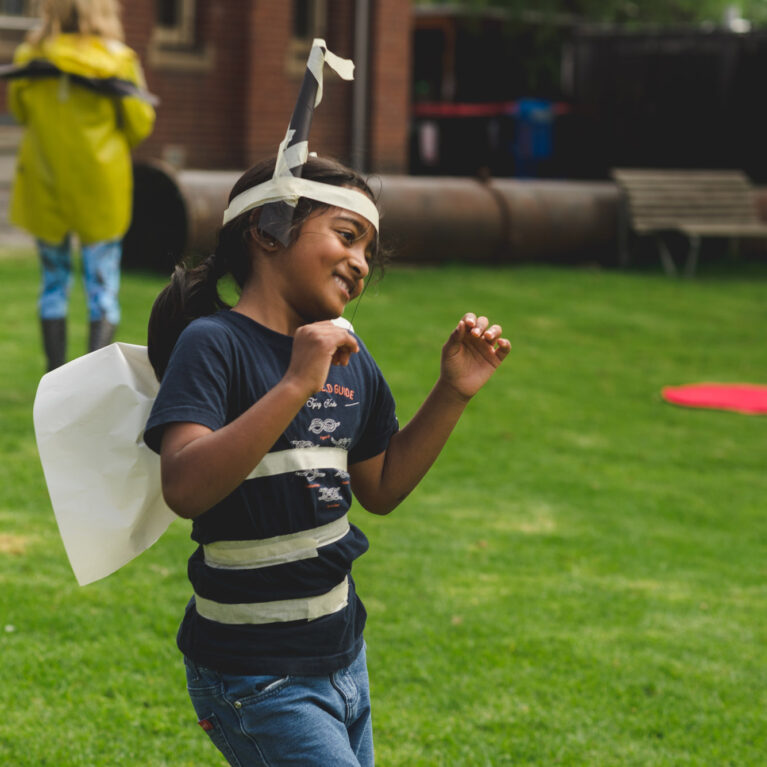 A Bees production photo. A child in a handmade paper and masking tape costume is smiling and copying the movements of the Bee (off camera) that they are watching. They are on a green lawn, with other people visible in the background.